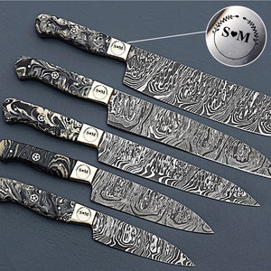 May include: A set of six Damascus steel kitchen knives. The blades have a distinctive wave pattern. The handles are black and white marbled. Each knife has a small, round emblem with the initials "S" and "M" inside a heart and wreath.