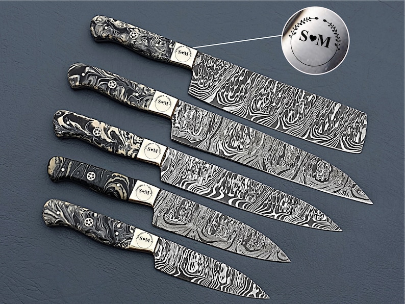 May include: A set of six Damascus steel kitchen knives. The blades have a distinctive wave pattern. The handles are black and white marbled. Each knife has a small, round emblem with the initials "S" and "M" inside a heart and wreath.