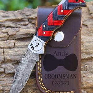 May include: A folding pocket knife with a Damascus steel blade and a red and black woven pattern handle. The metal bolster is engraved with A&B. It rests on a brown leather sheath featuring a bow tie icon, the name Andy, and GROOMSMAN 02-28-23.