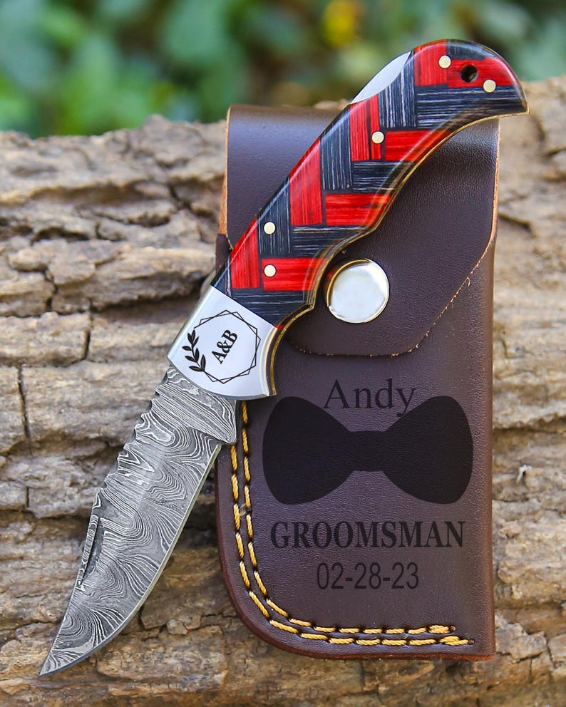 May include: A folding pocket knife with a Damascus steel blade and a red and black woven pattern handle. The metal bolster is engraved with A&B. It rests on a brown leather sheath featuring a bow tie icon, the name Andy, and GROOMSMAN 02-28-23.