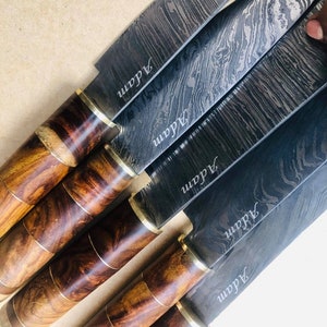 May include: A collection of Damascus steel knives with wooden handles. The blades have a unique swirling pattern and the name "Adam" is etched on each blade. The handles are a mix of dark and light wood tones, with brass accents.