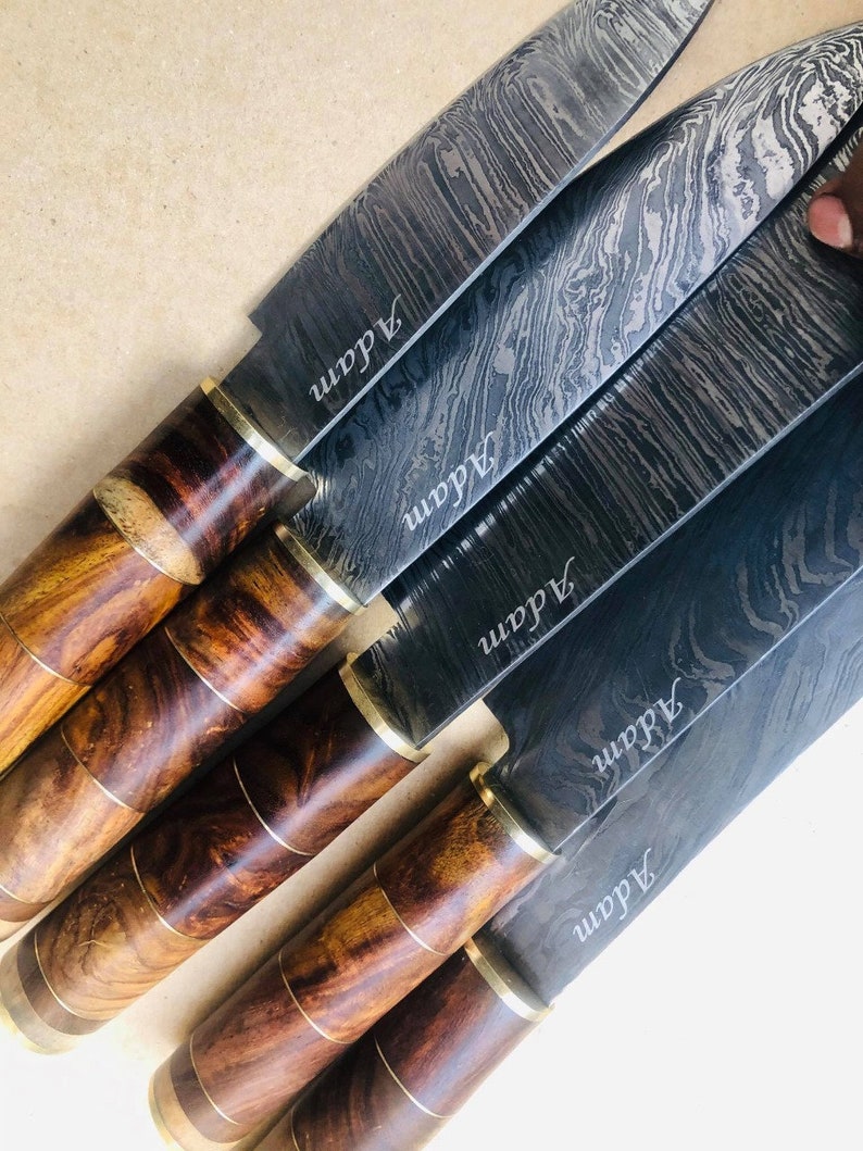 May include: A collection of Damascus steel knives with wooden handles. The blades have a unique swirling pattern and the name "Adam" is etched on each blade. The handles are a mix of dark and light wood tones, with brass accents.