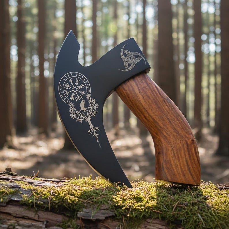 May include: A black axe with a wooden handle, featuring engraved silver-coloured Viking-inspired designs. The axe blade displays a tree and bird motif, and a circular symbol. The handle is made of dark wood.