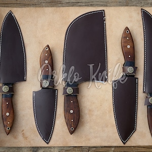 May include: A set of five kitchen knives with dark brown wooden handles and matching leather sheaths. The knives are displayed on a tan leather roll-up case with buckle straps. The blades are various shapes and sizes.