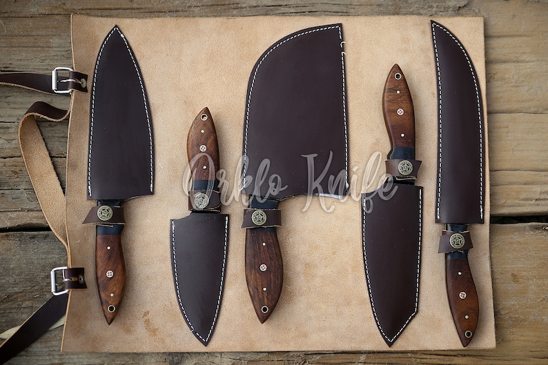 May include: A set of five kitchen knives with dark brown wooden handles and matching leather sheaths. The knives are displayed on a tan leather roll-up case with buckle straps. The blades are various shapes and sizes.