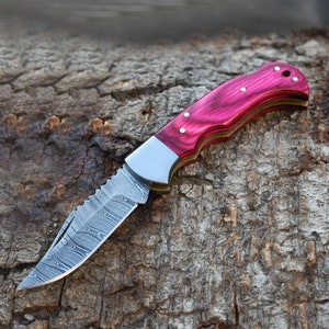 May include: A folding pocket knife featuring a silver Damascus steel blade with a distinct wavy pattern, a metallic bolster, and a curved handle made of polished magenta wood with three visible brass pins and a lanyard hole at the end.