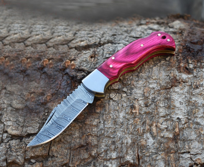 May include: A folding pocket knife featuring a silver Damascus steel blade with a distinct wavy pattern, a metallic bolster, and a curved handle made of polished magenta wood with three visible brass pins and a lanyard hole at the end.