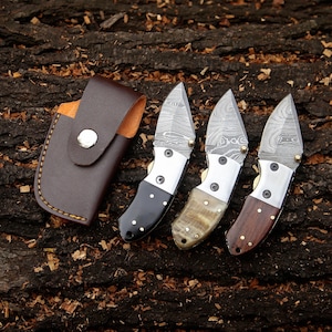 May include: Three small folding knives with patterned blades and various handle materials, including black, tan, and brown wood. A brown leather sheath with a snap closure is also present. The knives are displayed on a textured wood surface.