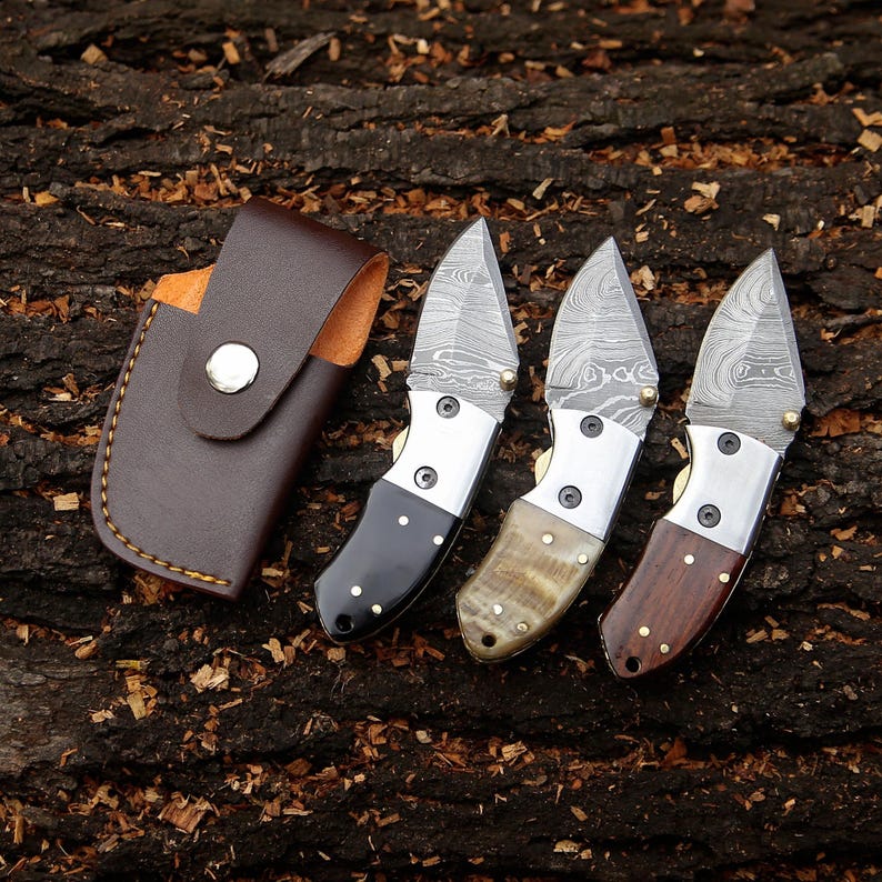 May include: Three small folding knives with patterned blades and various handle materials, including black, tan, and brown wood. A brown leather sheath with a snap closure is also present. The knives are displayed on a textured wood surface.