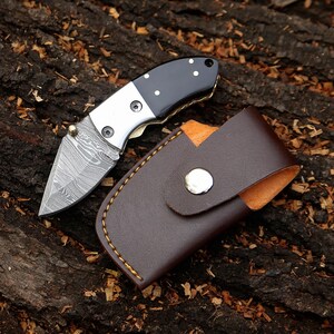May include: A small, folding pocket knife with a patterned blade and a black and silver handle. The knife is shown with a brown leather sheath. The blade has a unique, textured pattern. The sheath has a snap closure.