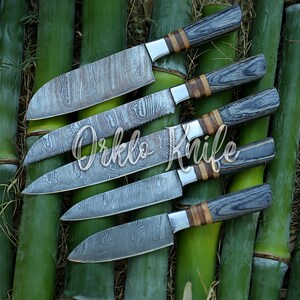 May include: An assortment of five kitchen knives with patterned blades and wooden handles. The handles are a mix of gray and brown wood, with silver bolsters. The knives are arranged on a bed of green bamboo stalks. The text "Orklo Knife" is overlaid on the image.