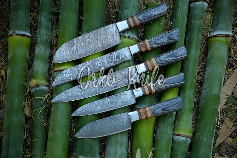 May include: An assortment of five kitchen knives with patterned blades and wooden handles. The handles are a mix of gray and brown wood, with silver bolsters. The knives are arranged on a bed of green bamboo stalks. The text "Orklo Knife" is overlaid on the image.