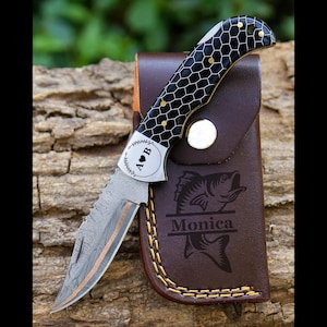 May include: A folding knife with a patterned blade and a black and gold handle with a honeycomb design. The knife is in a brown leather sheath with a fish graphic and the name "Monica". The blade has a wavy edge.