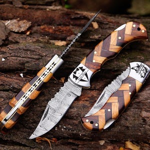 May include: Three folding knives with handles made of light and dark wood in a herringbone pattern. One knife is open, showing a Damascus steel blade with a Best Man tuxedo graphic. Another is closed with a fish graphic and the name Monica.
