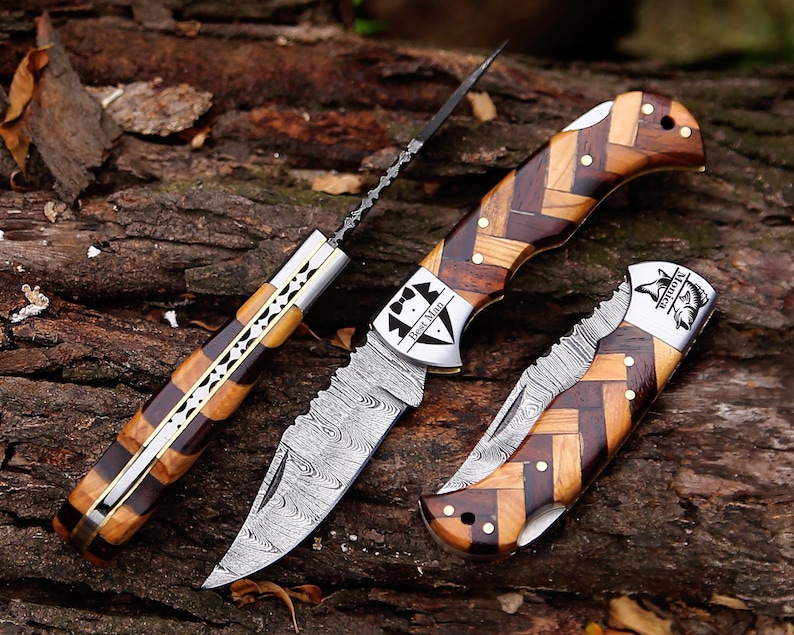 May include: Three folding knives with handles made of light and dark wood in a herringbone pattern. One knife is open, showing a Damascus steel blade with a Best Man tuxedo graphic. Another is closed with a fish graphic and the name Monica.