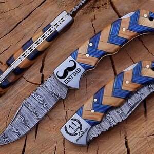 May include: Three folding knives with patterned blades and handles. The handles feature a blue and wood grain design. One knife has "BEST DAD" and a mustache graphic. Another has "11 YEARS ANNIVERSARY".
