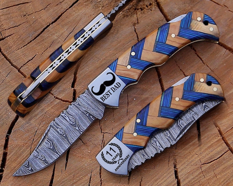 May include: Three folding knives with patterned blades and handles. The handles feature a blue and wood grain design. One knife has "BEST DAD" and a mustache graphic. Another has "11 YEARS ANNIVERSARY".