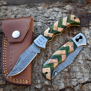 May include: Two folding pocket knives with Damascus steel blades and handles featuring a woven pattern of natural wood and green segments. One blade is open, one is closed. The open blade is engraved with "LY Love You". The closed blade has a bow tie icon, "Name", "GROOMSMAN", and "02-28-23". A brown leather sheath is included.