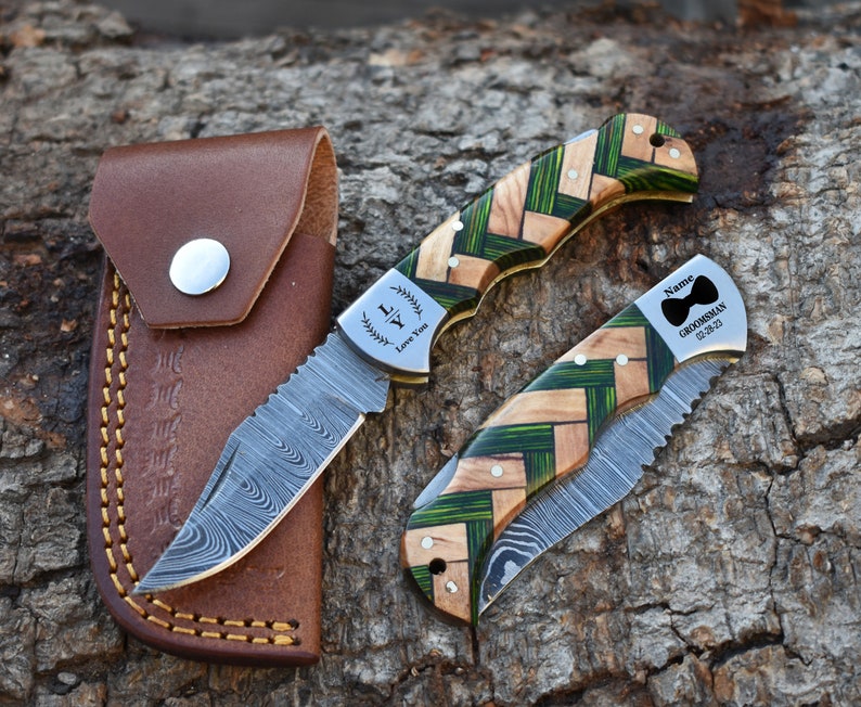 May include: Two folding pocket knives with Damascus steel blades and handles featuring a woven pattern of natural wood and green segments. One blade is open, one is closed. The open blade is engraved with "LY Love You". The closed blade has a bow tie icon, "Name", "GROOMSMAN", and "02-28-23". A brown leather sheath is included.