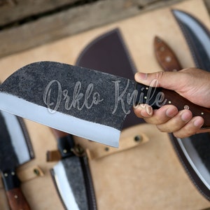 May include: A large, dark-colored cleaver knife with a wooden handle is held in a hand. The blade has a textured finish and a sharp edge. The text "Orklo Knife" is visible on the blade. Other knives are in the background.