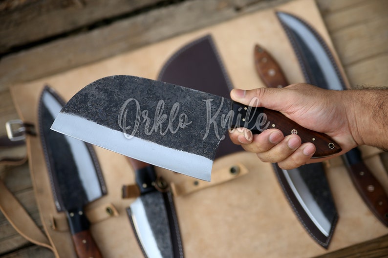 May include: A large, dark-colored cleaver knife with a wooden handle is held in a hand. The blade has a textured finish and a sharp edge. The text "Orklo Knife" is visible on the blade. Other knives are in the background.