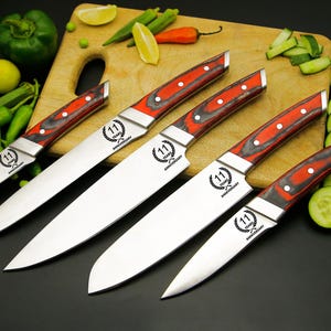 May include: A set of five stainless steel kitchen knives with red and black wooden handles. The blades are arranged on a wooden cutting board with vegetables and fruit. Each knife has the text "11 YEARS ANNIVERSARY".