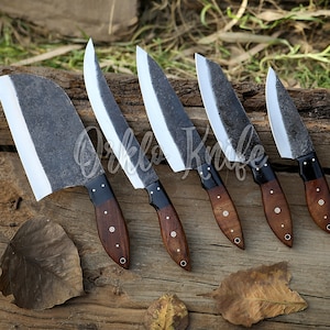 May include: Five handcrafted knives with dark, textured blades and wooden handles are arranged on a wooden surface. The blades have a bright, polished edge. The handles are dark brown with decorative rivets. The text "Orklo Knife" is visible.