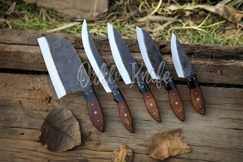 May include: Five handcrafted knives with dark, textured blades and wooden handles are arranged on a wooden surface. The blades have a bright, polished edge. The handles are dark brown with decorative rivets. The text "Orklo Knife" is visible.