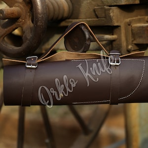 May include: A dark brown leather knife roll with a handle, secured by two straps with silver buckles. The roll has white stitching and the text "Orklo Knife" is visible. The background features a rusty metal wheel.
