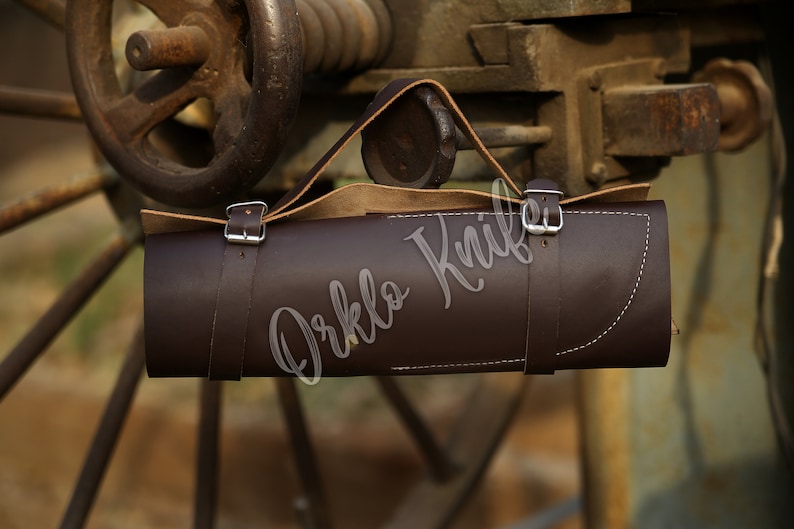 May include: A dark brown leather knife roll with a handle, secured by two straps with silver buckles. The roll has white stitching and the text "Orklo Knife" is visible. The background features a rusty metal wheel.