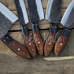 May include: Five handmade cleaver knives with dark, textured blades and wooden handles. The handles are a rich brown with black accents and decorative rivets. The knives are arranged on a rustic wooden surface.