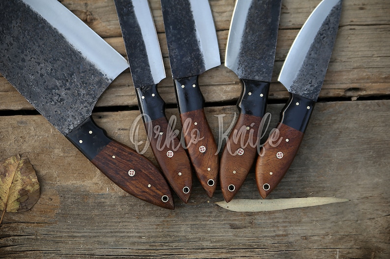 May include: Five handmade cleaver knives with dark, textured blades and wooden handles. The handles are a rich brown with black accents and decorative rivets. The knives are arranged on a rustic wooden surface.