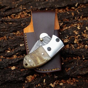 May include: A folding pocket knife with a patterned blade and a horn handle, resting in a brown leather sheath. The knife has a silver bolster and is secured with small rivets. The sheath is stitched with yellow thread.