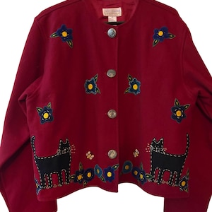 May include: A burgundy button-up jacket with embroidered details. The jacket features black cat and blue flower designs. Silver buttons and a round neckline complete the look. The jacket is hanging on a black hanger.