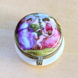May include: A round porcelain trinket box with a brass metal rim and hinge. The lid features a colorful painted scene of two figures in flowing purple and pink robes sitting in a green landscape with a small child reaching for hanging grapes.