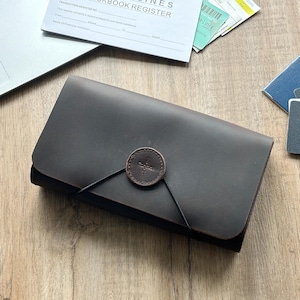May include: A dark brown leather wallet with a circular button closure and a matching leather strap. The wallet is on a wooden surface, with a laptop, checkbook register, passport, and cards nearby.