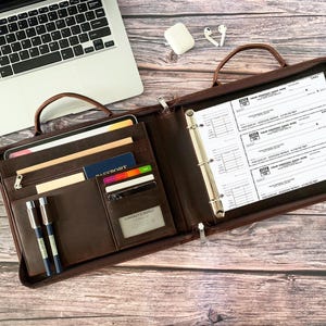 May include: Open brown leather business portfolio with a handle, zipper, and multiple compartments. Inside are pens, cards, a passport, and a tablet. Checks are visible in the binder section. A laptop, earbuds, and a white charging case are in the background.