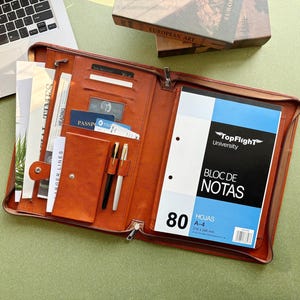 May include: Open brown leather portfolio with a zipper closure, containing a notepad, pens, and documents. The notepad has a blue and white cover with the text "BLOC DE NOTAS". A laptop and a book titled "EUROPEAN ART" are in the background.