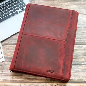 May include: A burgundy leather portfolio with a zipper closure, a stitched pocket, and the embossed initials "MERO M." A laptop and a card with "Best Wishes For you" are also in the image. The portfolio is on a wooden surface.