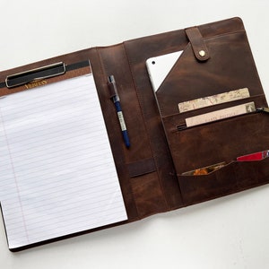 May include: A brown leather portfolio with a clipboard, pen, and multiple pockets. The clipboard has lined paper and the text "Verilus" printed on it.