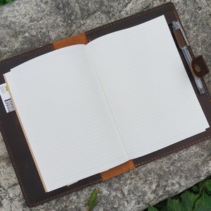 May include: A brown leather journal with a pen holder and a pocket on the side. The journal is open to a blank page with lined paper.