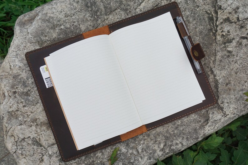 May include: A brown leather journal with a pen holder and a pocket on the side. The journal is open to a blank page with lined paper.
