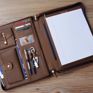 May include: A brown leather portfolio with a zipper closure, multiple pockets, and a notepad. The portfolio is open and has a pen holder, a card slot, and a phone pocket.