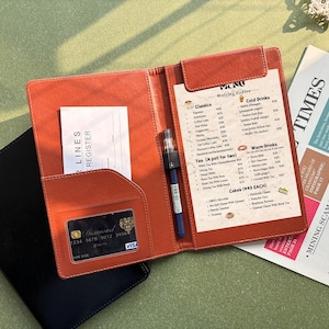 May include: An open, tan leather menu holder with a menu, pen, and receipt. The menu lists coffee, tea, and cakes. A black credit card is visible in a pocket. A newspaper is in the background.