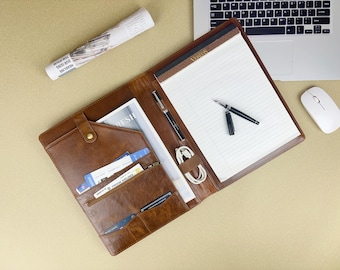Könnte beinhalten: Ein braunes Leder-Portfolio mit Notizblock, Stiften und Ladekabel. Das offene Portfolio zeigt Taschen mit Dokumenten und Karten. Ein Laptop und eine Maus sind im Hintergrund zu sehen. Eine zusammengerollte Zeitung liegt daneben.