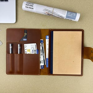 May include: A brown leather journal with pockets containing pens, cards, and a notebook. A rolled-up paper with the headline "DUR ISING TEM IPSUM" is on top, alongside a laptop and stationery.