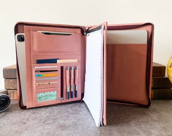 May include: A blush pink leather portfolio with a zipper closure. The portfolio is open to reveal a notepad, pen holders, card slots, and a pocket for a tablet or laptop.