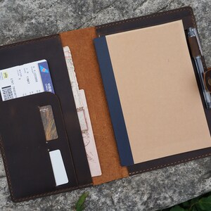 May include: A brown leather travel organizer with a pen holder, a pocket for a passport or boarding pass, and a notepad. The organizer is open and the notepad is blank.