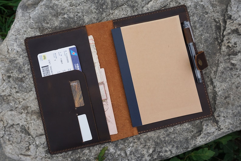 May include: A brown leather travel organizer with a pen holder, a pocket for a passport or boarding pass, and a notepad. The organizer is open and the notepad is blank.