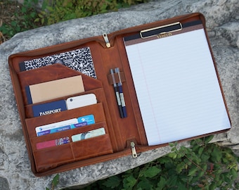 May include: A brown leather zippered portfolio with a notepad, pen holders, and multiple pockets for cards and a passport. The portfolio is open and lying on a rock.